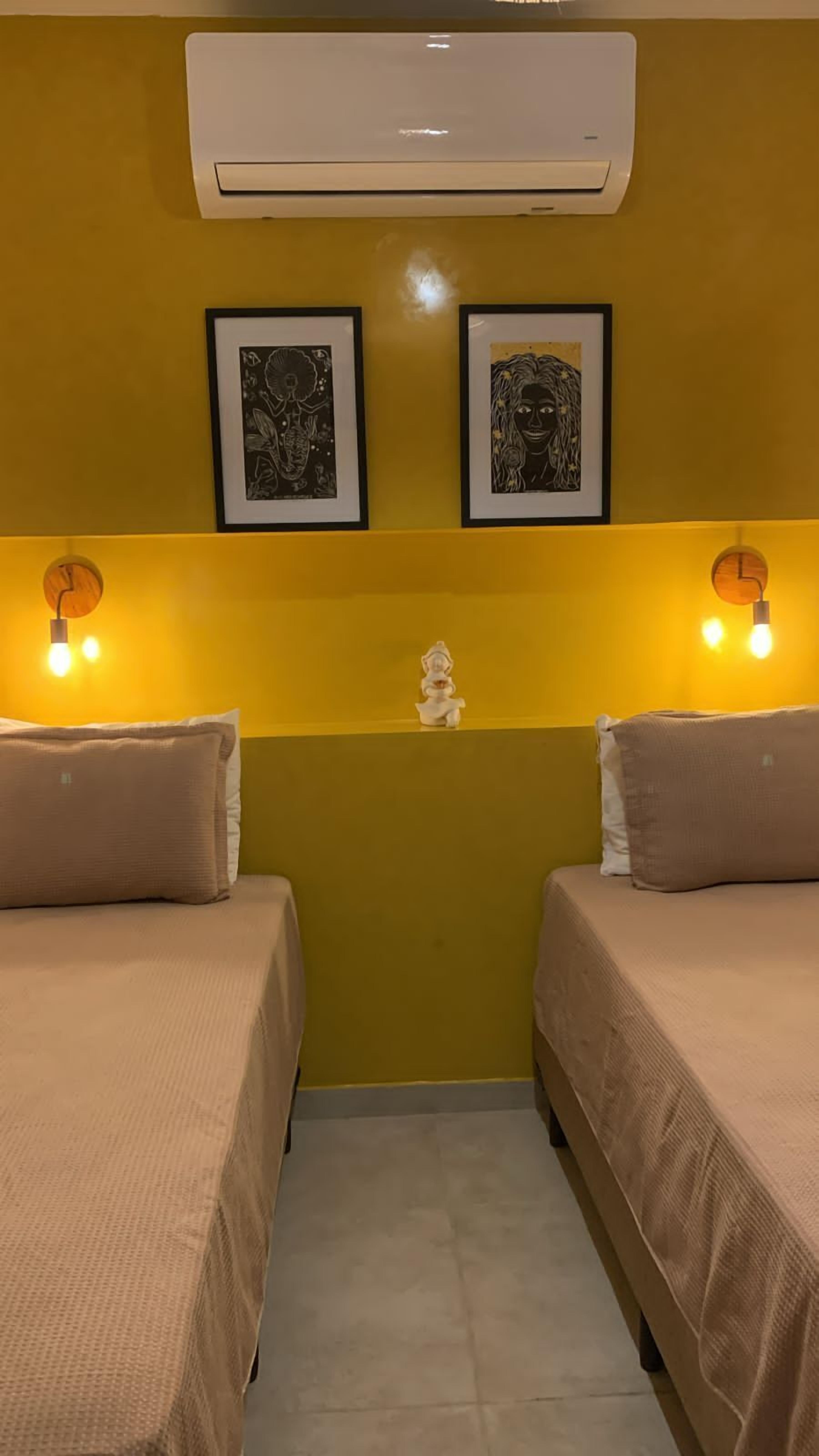 Una habitación con dos camas, aire acondicionado empotrado, dos cuadros enmarcados, una pequeña estatua e iluminación en la pared.