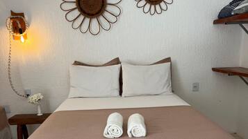 Una cama bien hecha con una colcha rosa, dos almohadas blancas y dos toallas enrolladas. Se observan dos elementos decorativos en la pared con forma de flores y una lámpara también en la pared.
