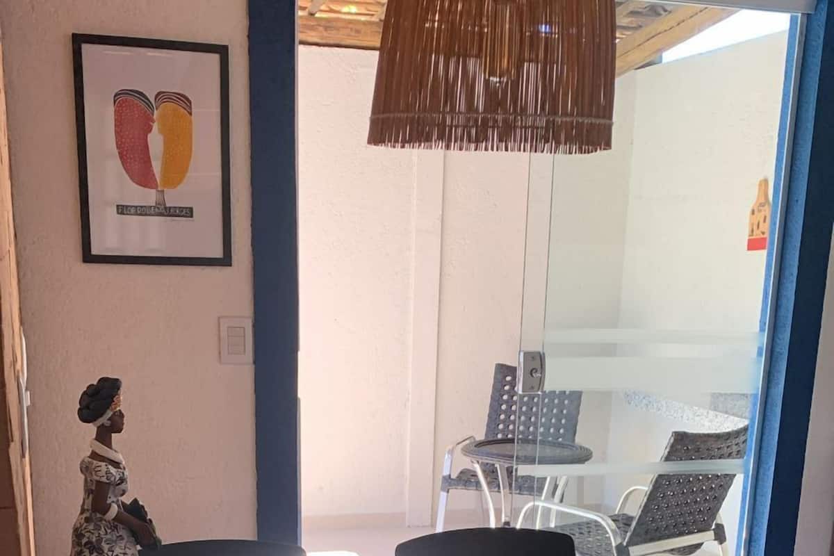 Un comedor con una mesa de vidrio y dos sillas negras. Se observa una obra de arte enmarcada en la pared y una lámpara colgante sobre la mesa.