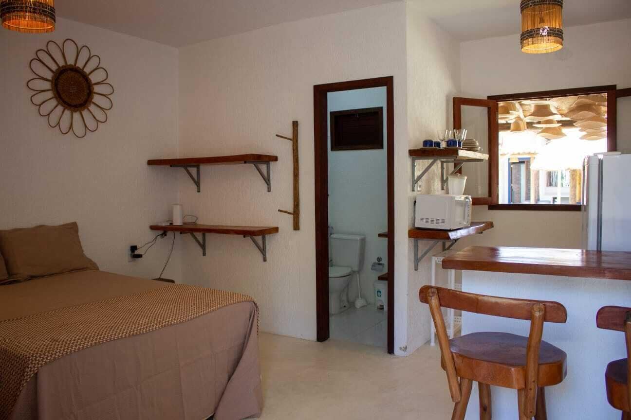 Une petite pièce comprenant un lit, une chaise en bois, une petite table et un miroir. Il y a des étagères au-dessus du lit et une porte donnant sur une salle de bain.