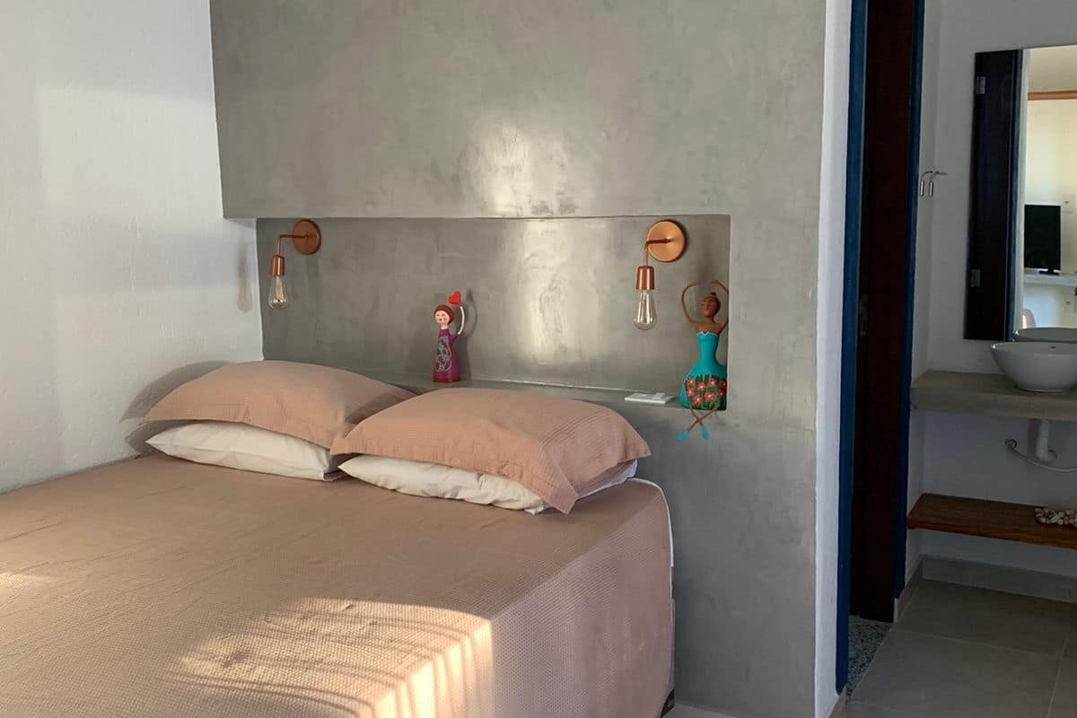Un dormitorio con cama, almohadas, cabecera, lámpara de pared y baño visible a través de una puerta abierta.