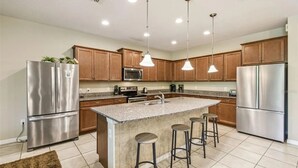 A modern kitchen with stainless steel appliances, granite countertops, wooden cabinets, and pendant lighting. - 9BR Home w/ Private Pool, Game Room, & Free Resort Access (Kissimmee)