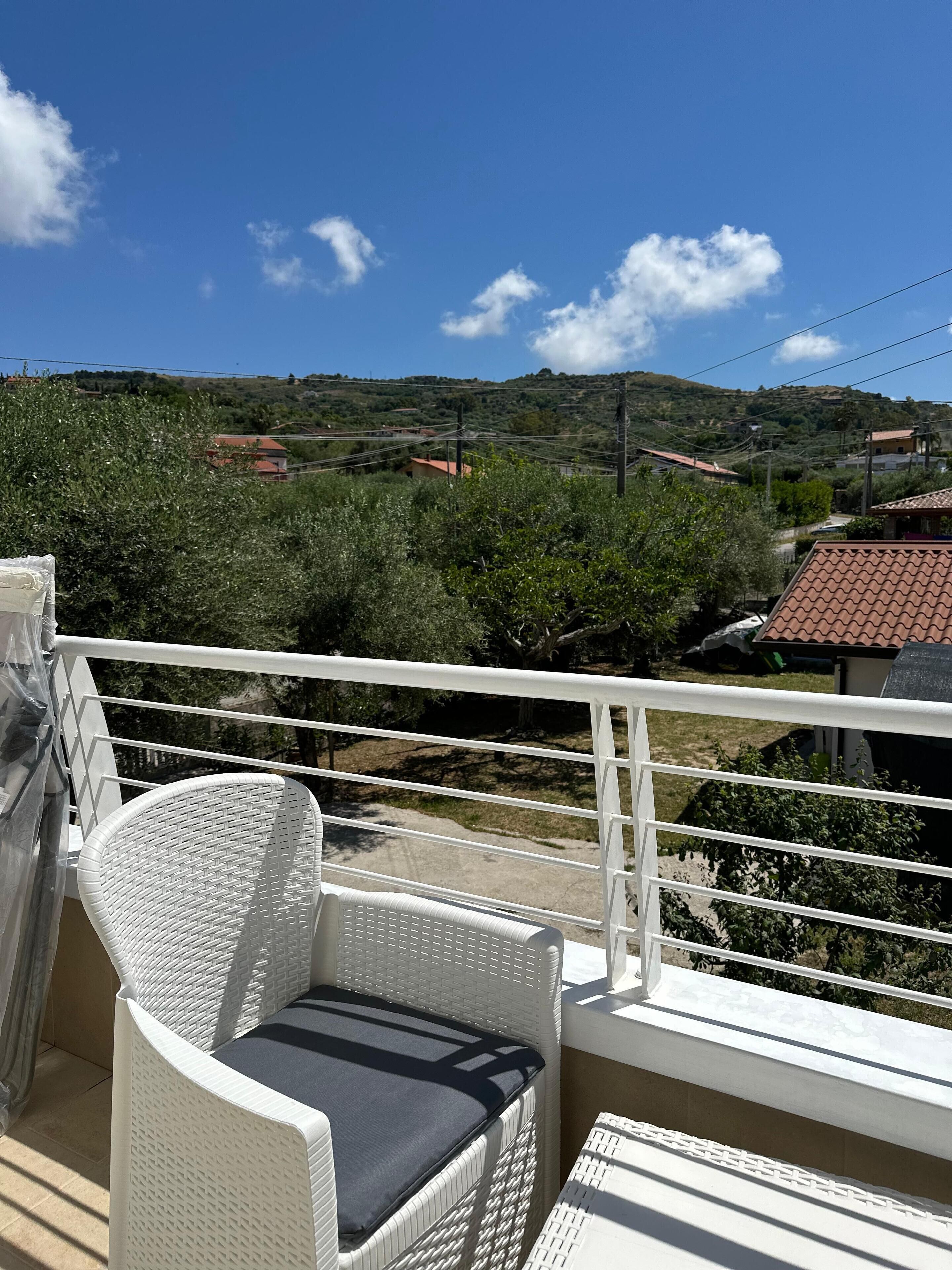 Un balcon avec une chaise en osier blanche et une petite table, donnant sur un paysage vallonné parsemé de maisons et de verdure.