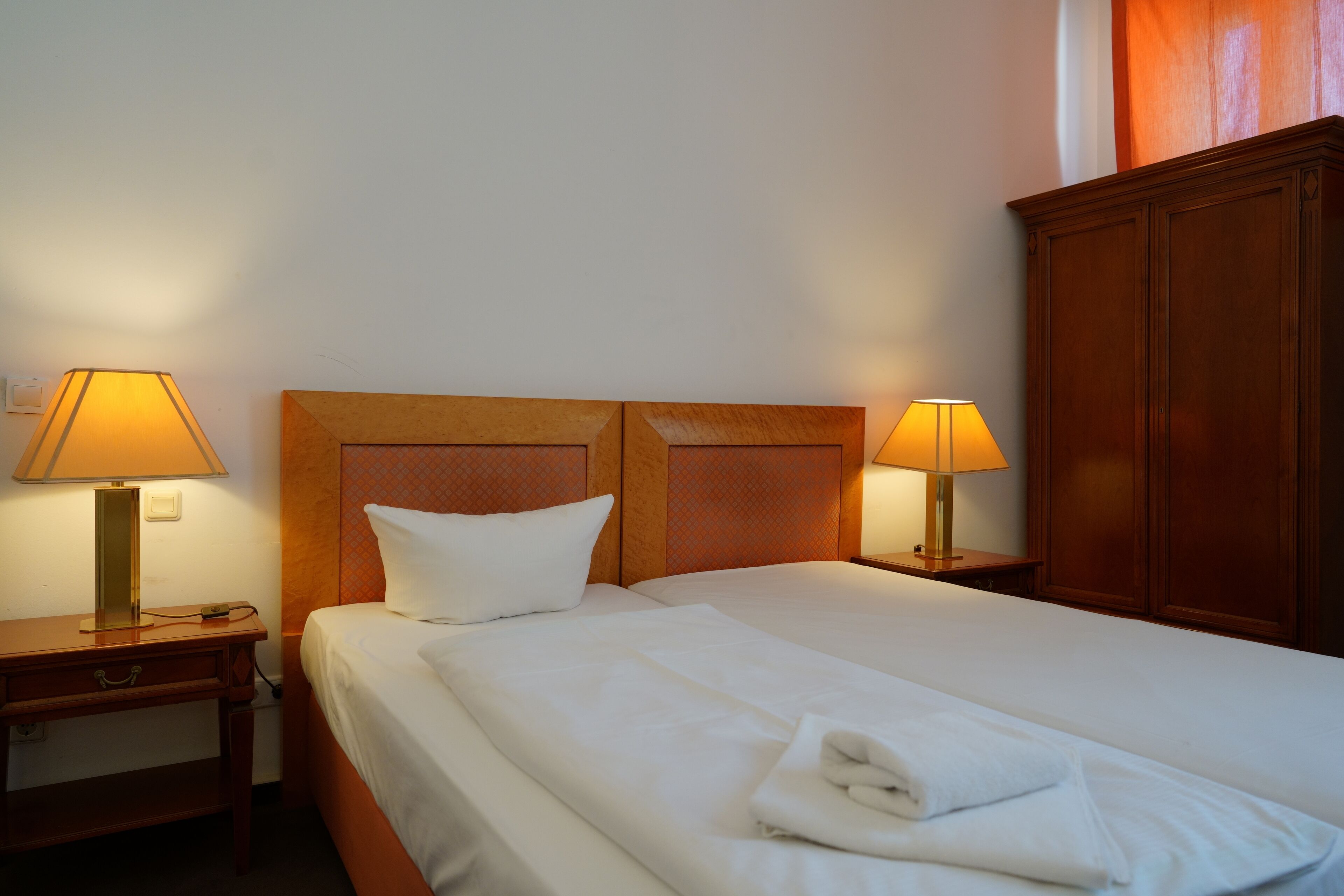 Una habitación de hotel con una cama bien hecha, dos lámparas de noche, un cabecero de madera y un armario de madera.