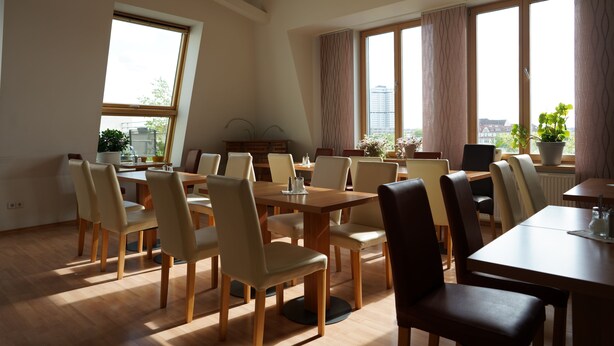 A spacious dining area with multiple tables and chairs, large windows with curtains, and potted plants. - Hotel Ambert (Berlin)