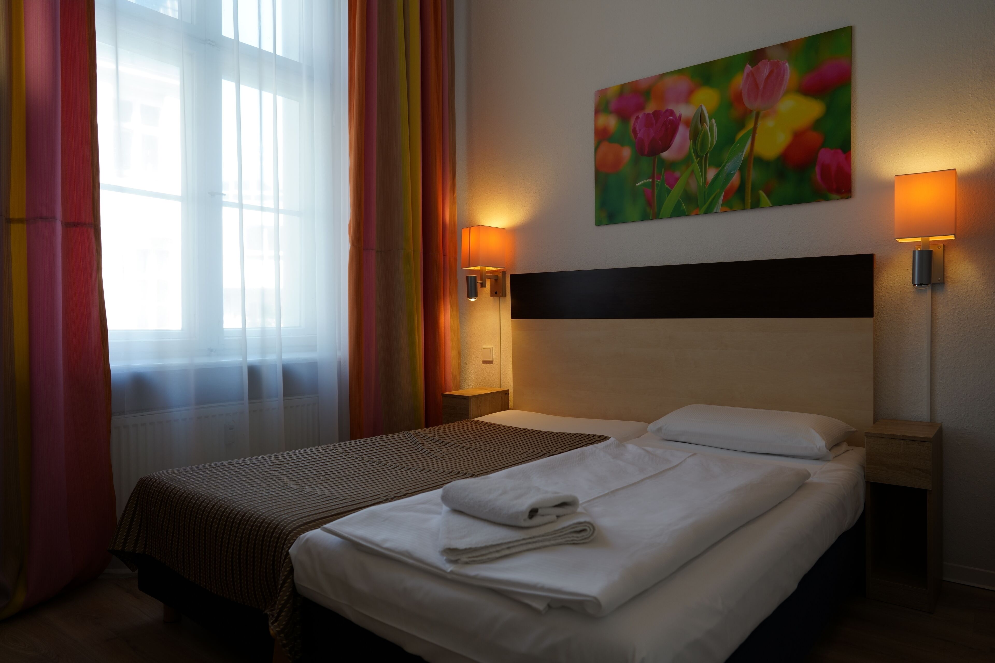 Et hotelværelse med en stor seng, to sengeborde, et blomsterbillede på væggen og et vindue med gardiner.