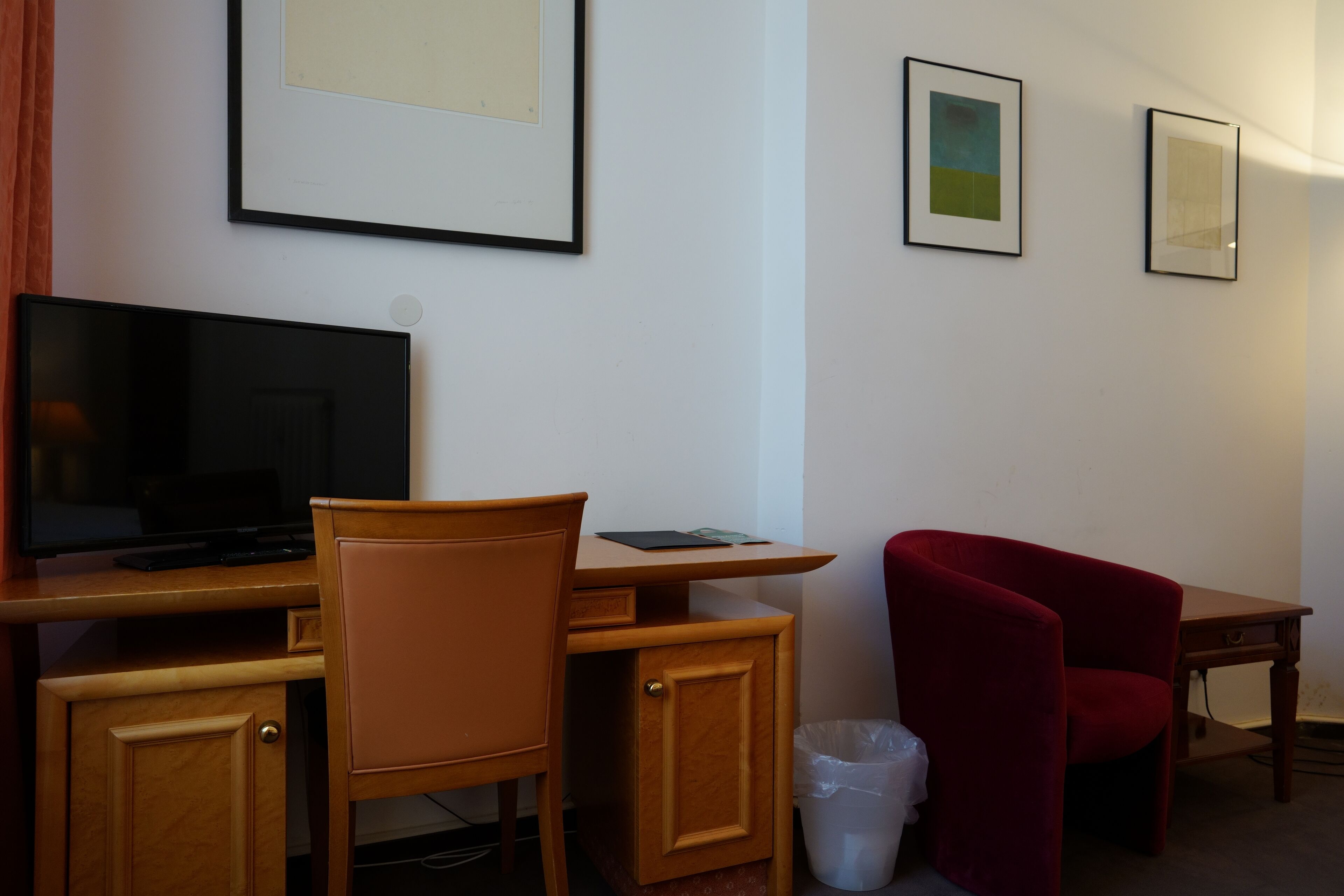 A hotel room with a wooden desk, a television, a red chair, and framed pictures on the wall.
