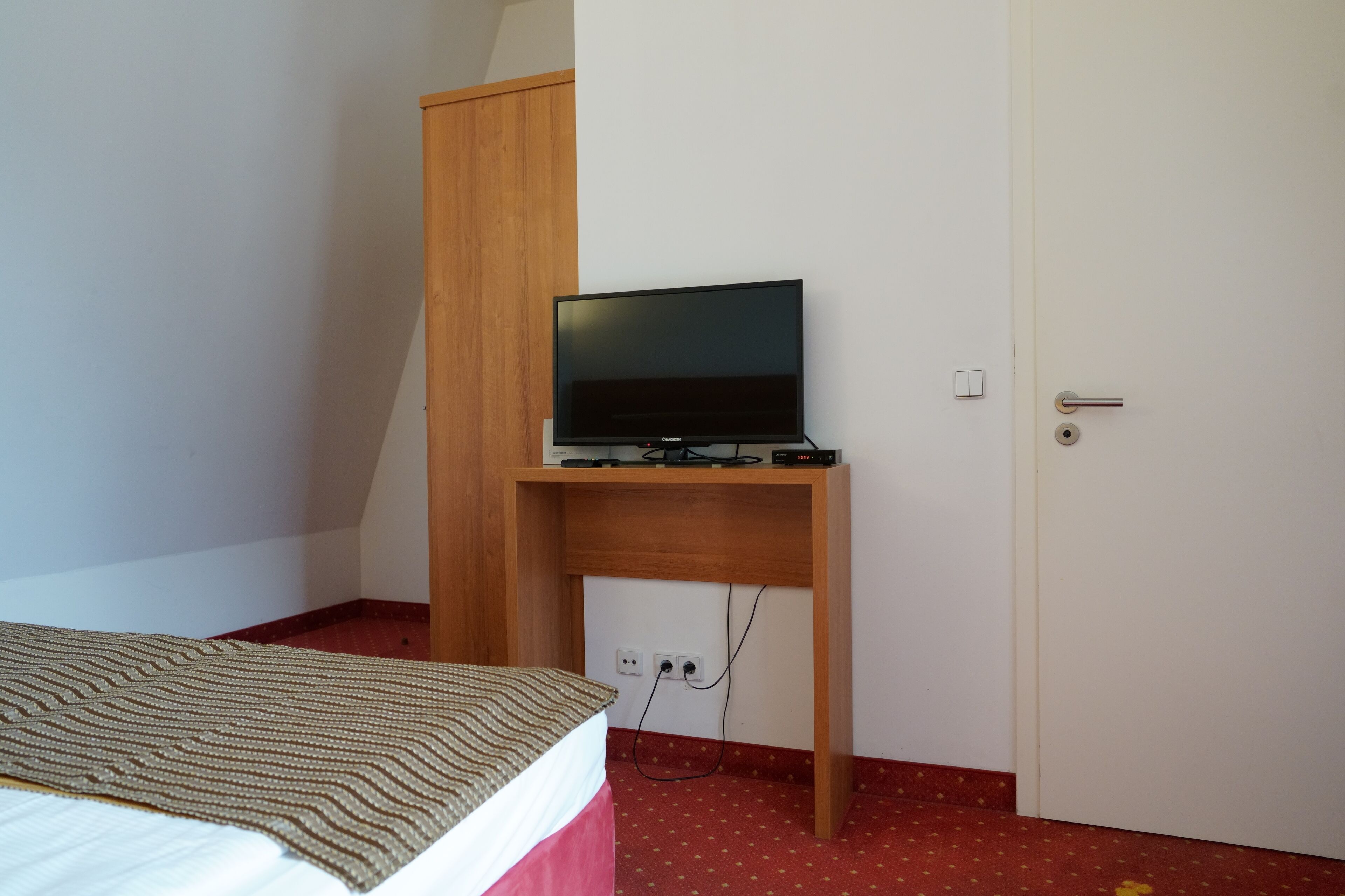 Een slaapkamer met een bed, een houten bureau met een televisie, een deur en een stopcontact.
