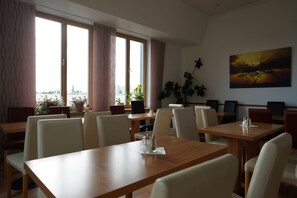 A dining area with tables and chairs, large windows with curtains, a potted plant, and a painting on the wall. - Hotel Ambert (Berlin)