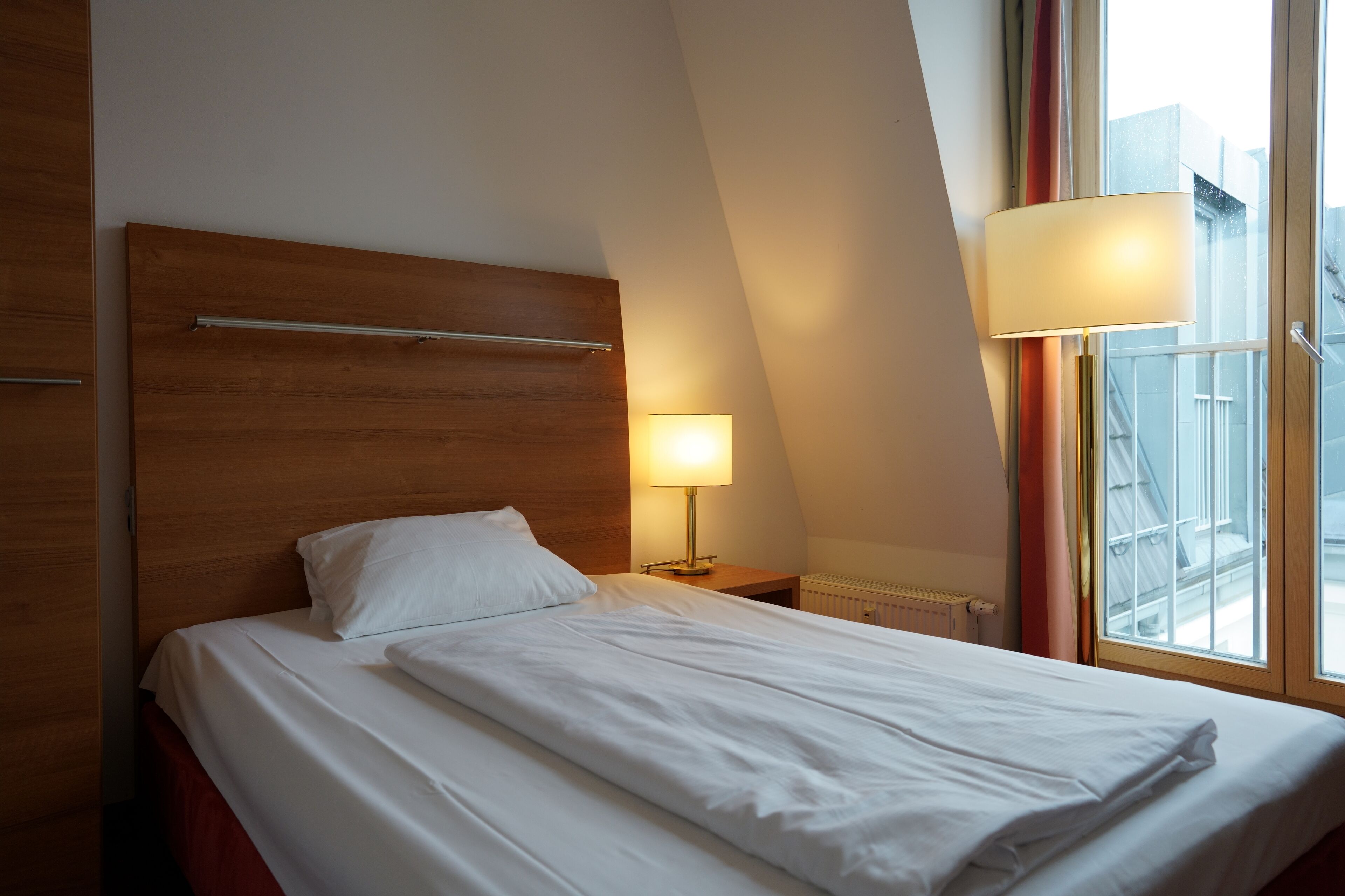 A hotel room with a wooden headboard, a single bed with white bedding, a bedside lamp, and a window with a view of buildings.