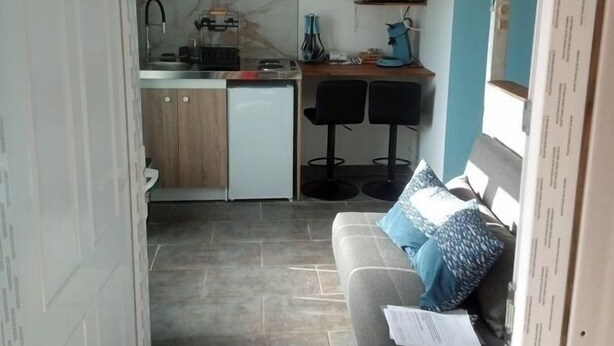 Una cocina compacta con lavamanos, estufa y microondas. Incluye un refrigerador pequeño, una zona de comedor con dos sillas y un sofá con cojín.