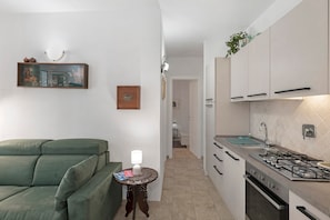 Eine moderne Küche mit weißen Schränken, einem Gasherd und einer Spüle. Ein grünes Sofa und ein kleiner runder Tisch mit einer Lampe sind ebenfalls vorhanden.