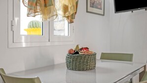 A dining table with a woven basket containing fruits, a window with a view of a cactus, and a television mounted on the wall.