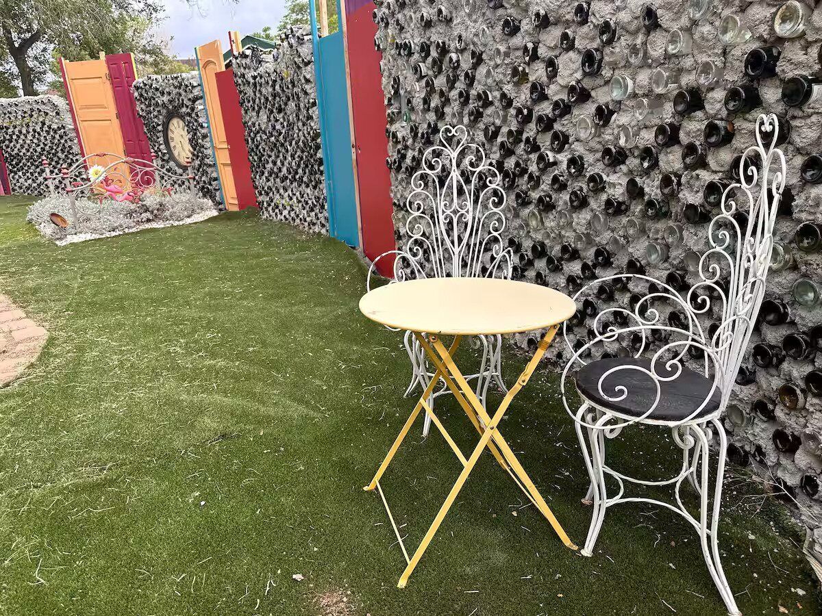 A small outdoor seating area with a round table and two chairs, surrounded by a decorative wall made of glass bottles.