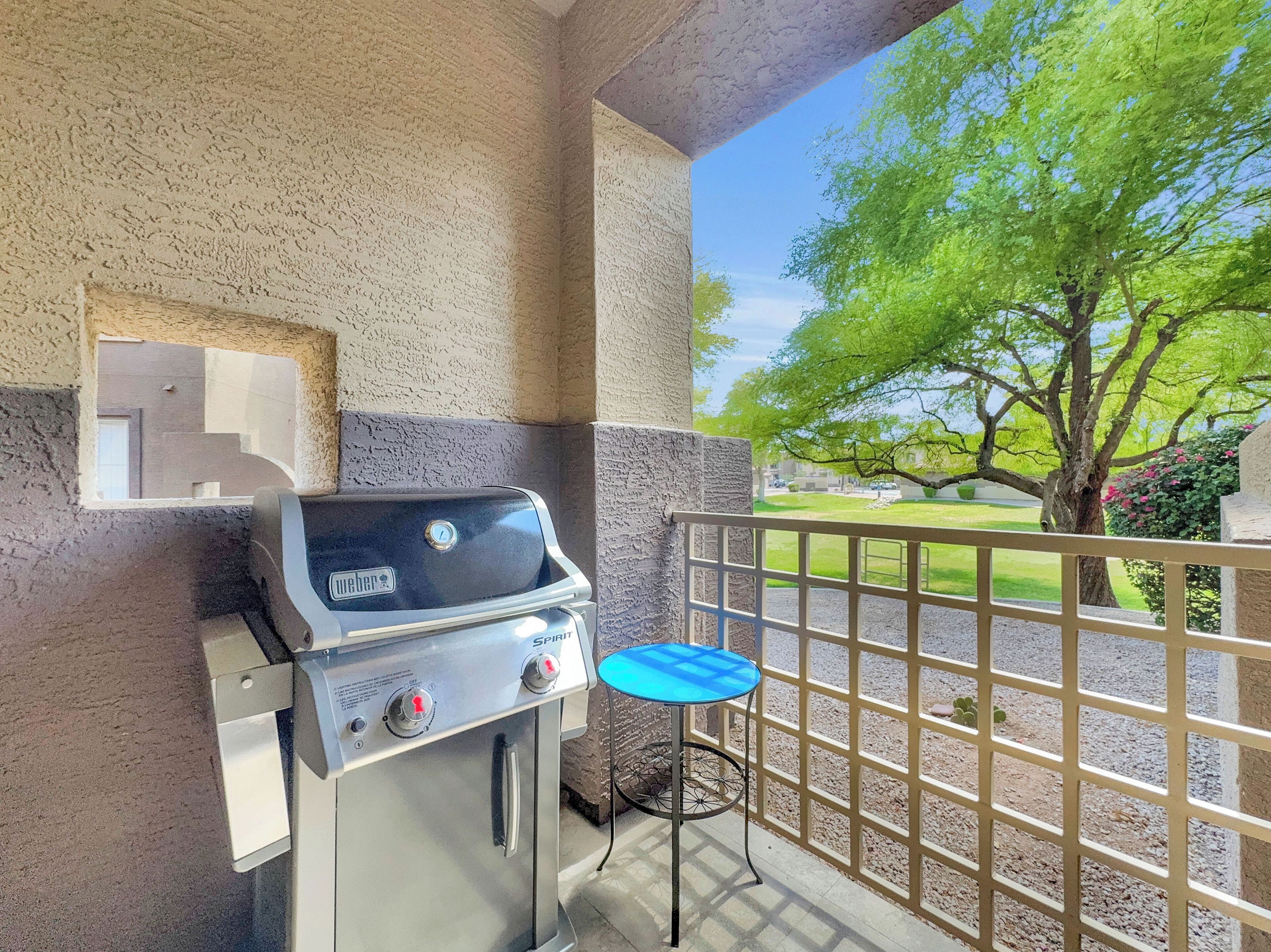 Una parrilla Weber Spirit junto a una mesa azul, frente a una pared con textura, con vista a un árbol verde y un área cercada.