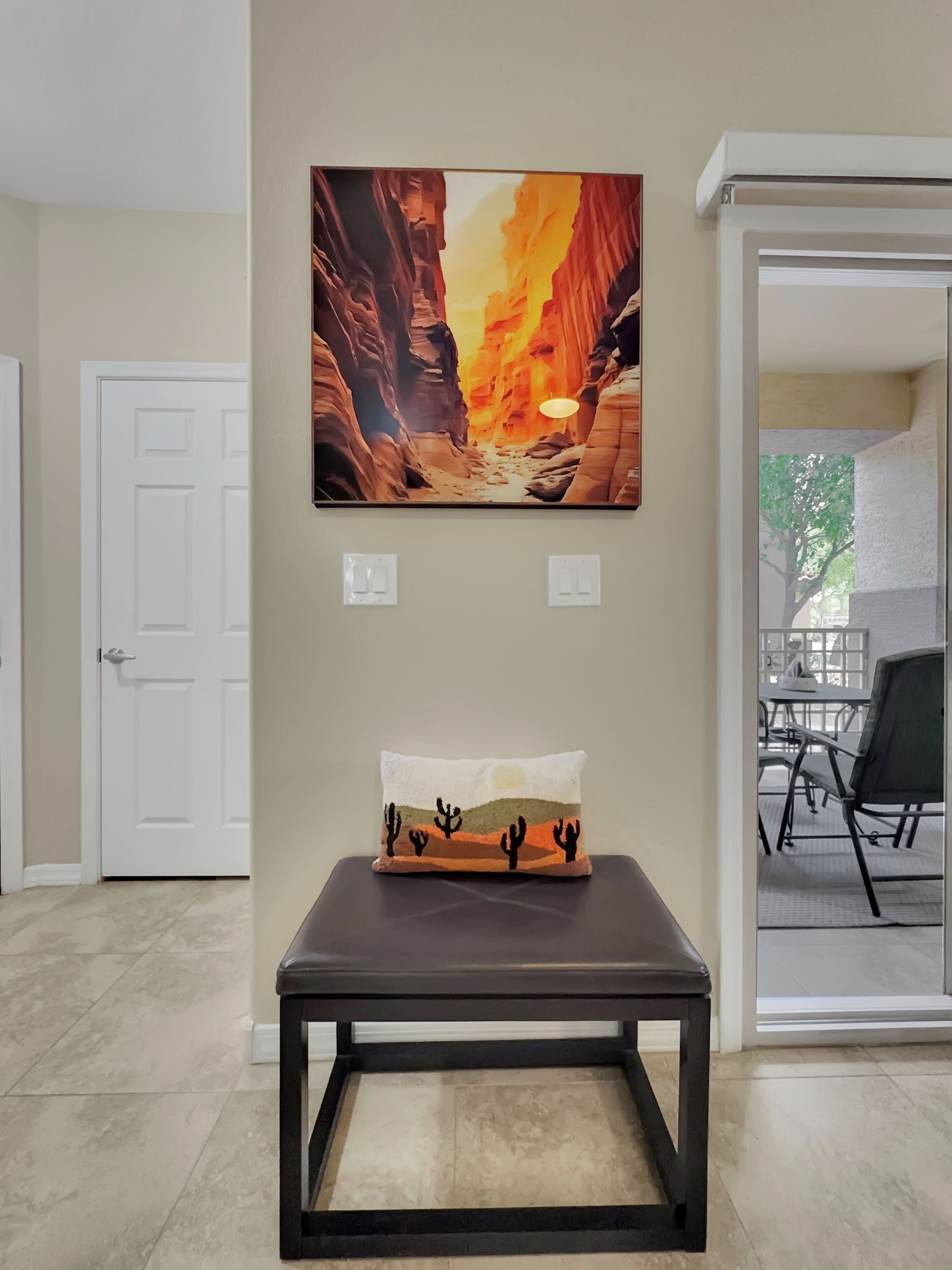 A room with a framed painting of a canyon, a small table with a cushion, and a door.