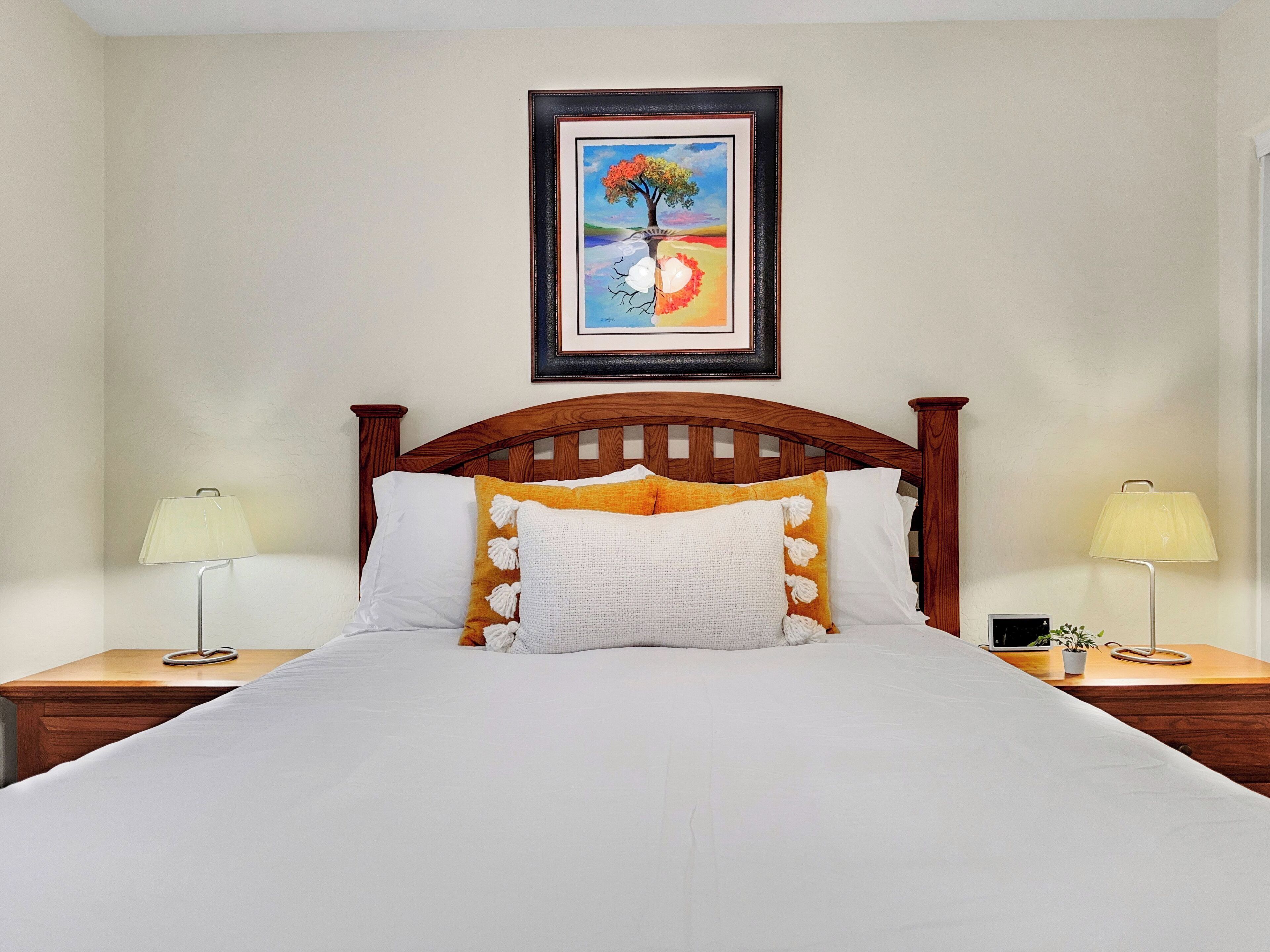 A neatly made bed with white and yellow pillows, a wooden headboard, and two bedside lamps. A framed painting hangs above the bed.