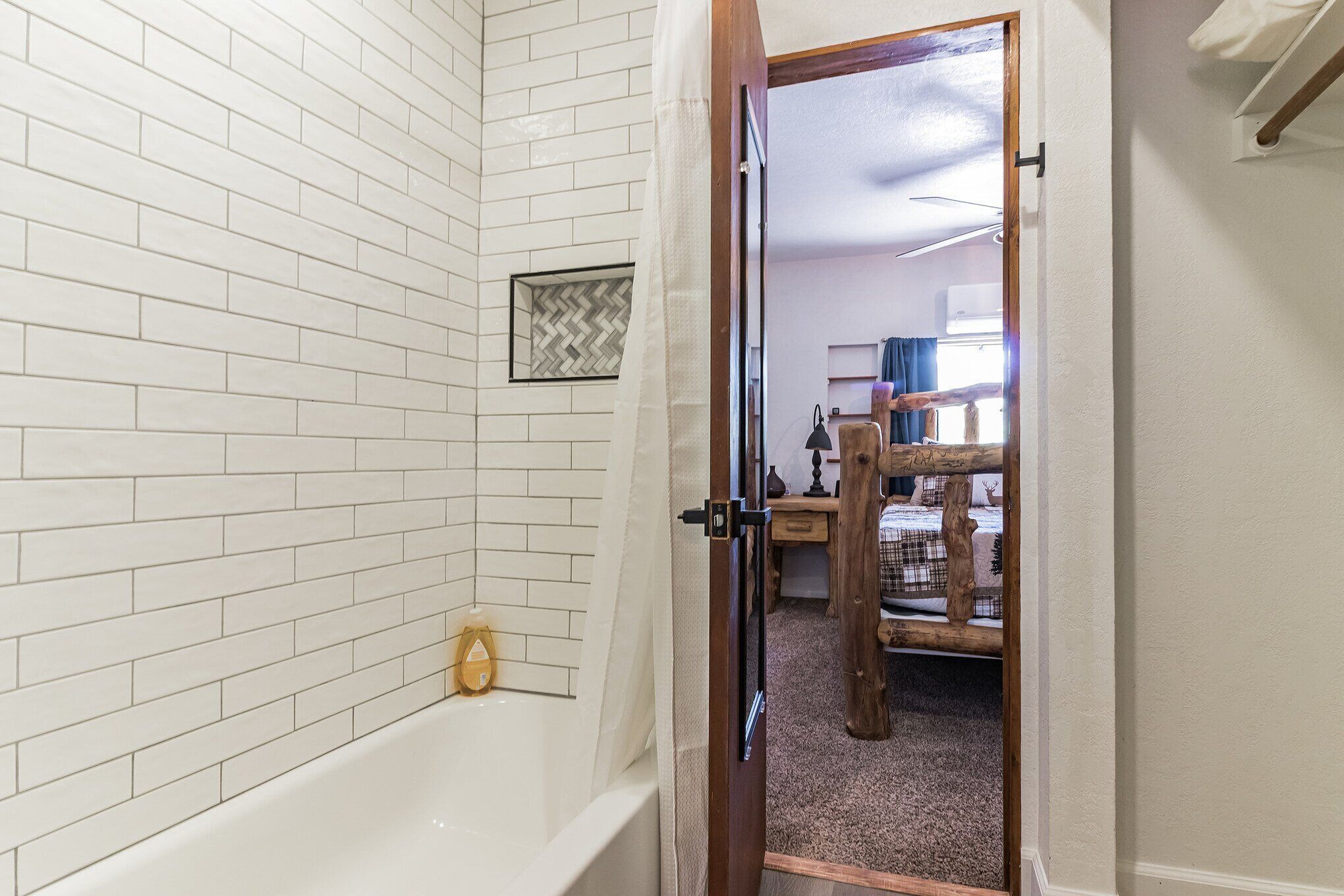 A bathroom with white tiled walls, a bathtub, and a wooden door leading to a bedroom with a bed and a dresser.