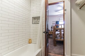A bathroom with white tiled walls, a bathtub, and a wooden door leading to a bedroom with a bed and a dresser. - Prince Pine (Pine)