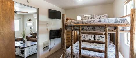 A bunk bed with a forest-themed mattress, a television mounted on the wall, and a wooden desk with a plant.