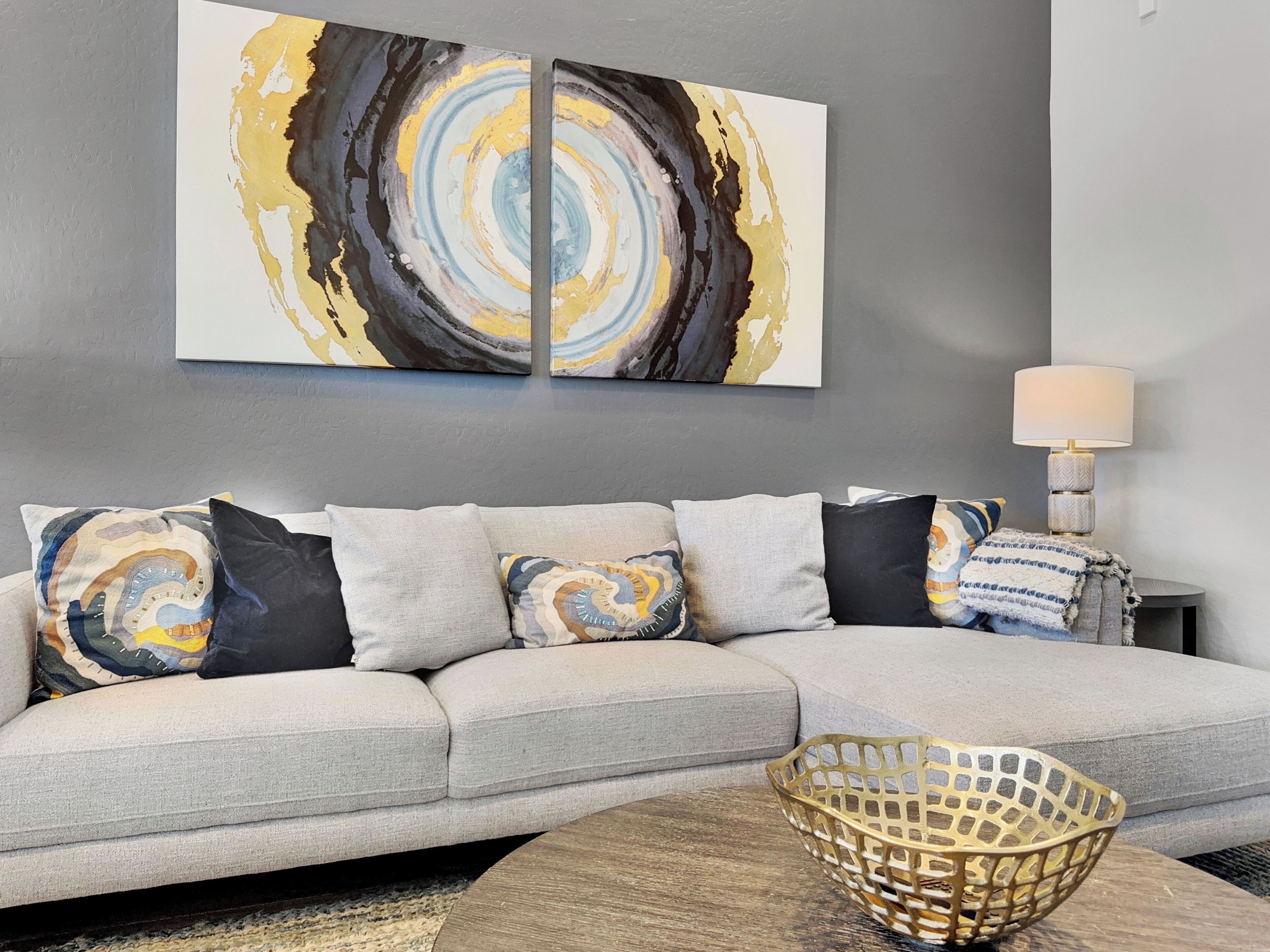A modern living room with a grey sofa, patterned cushions, a gold woven side table, and a large abstract painting on the wall.