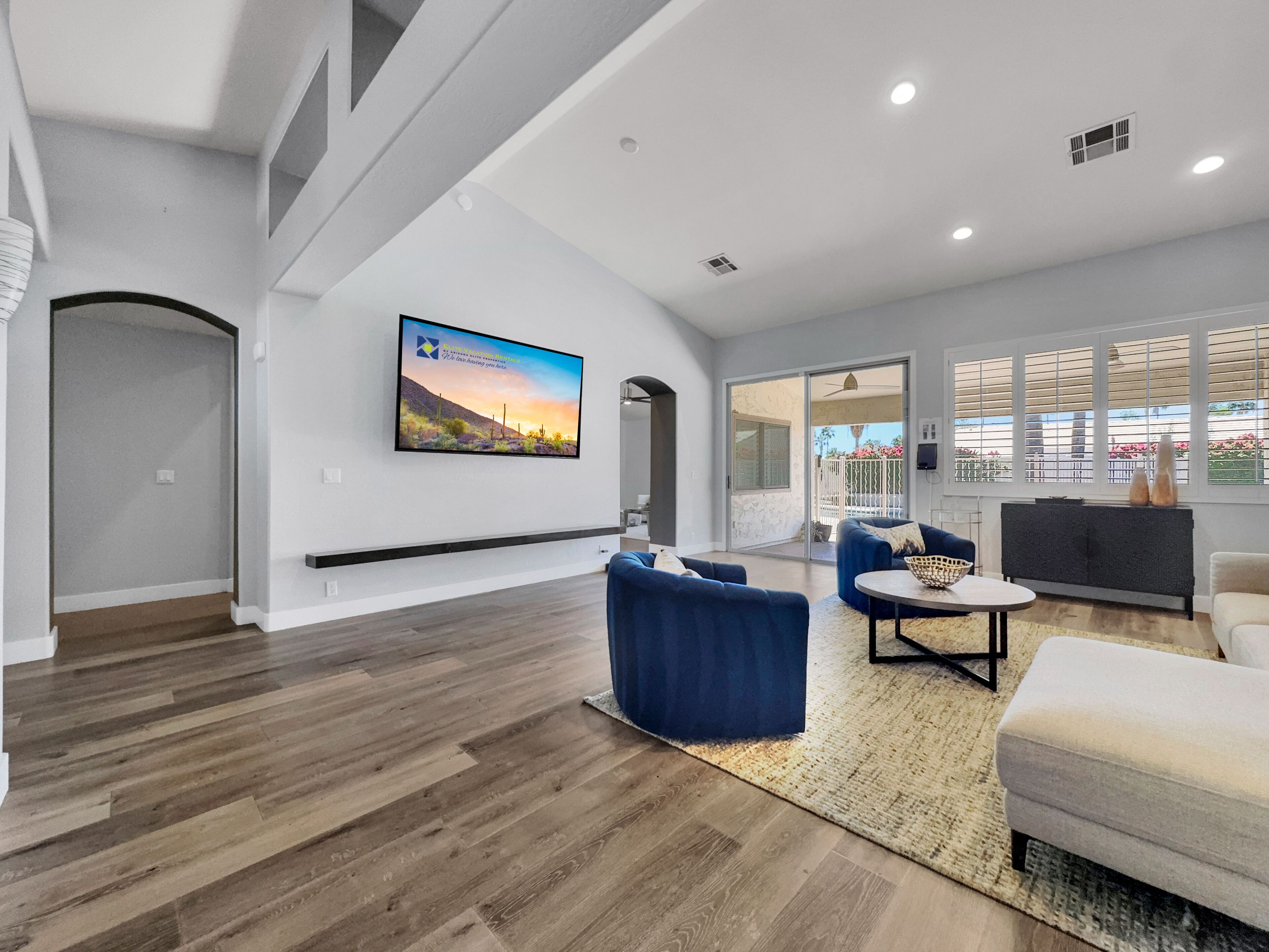 A spacious living room with a large flat-screen TV mounted on the wall, a blue sofa, a white armchair, and a small coffee table.
