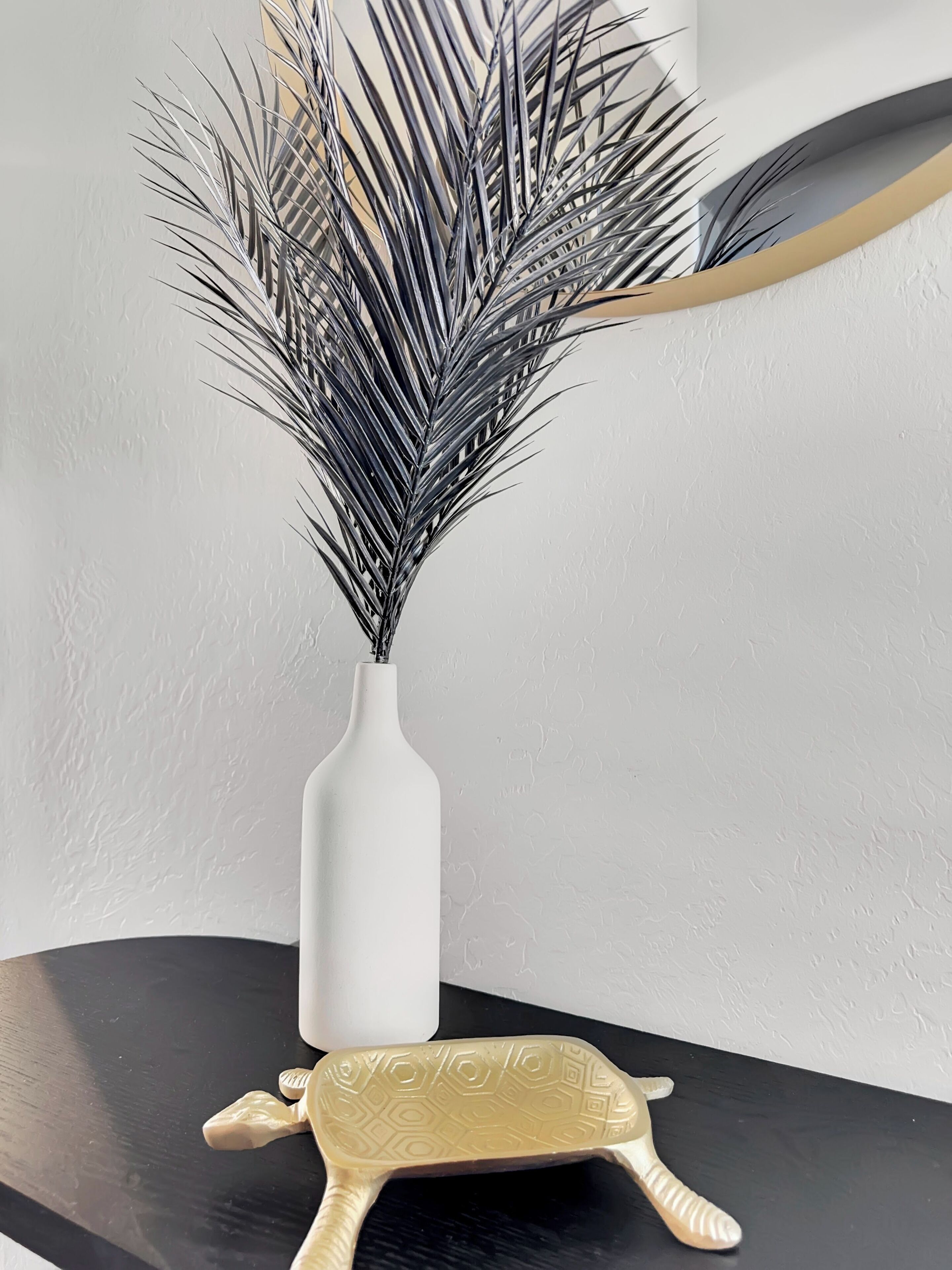 A white vase with a plant on a black table against a textured wall.
