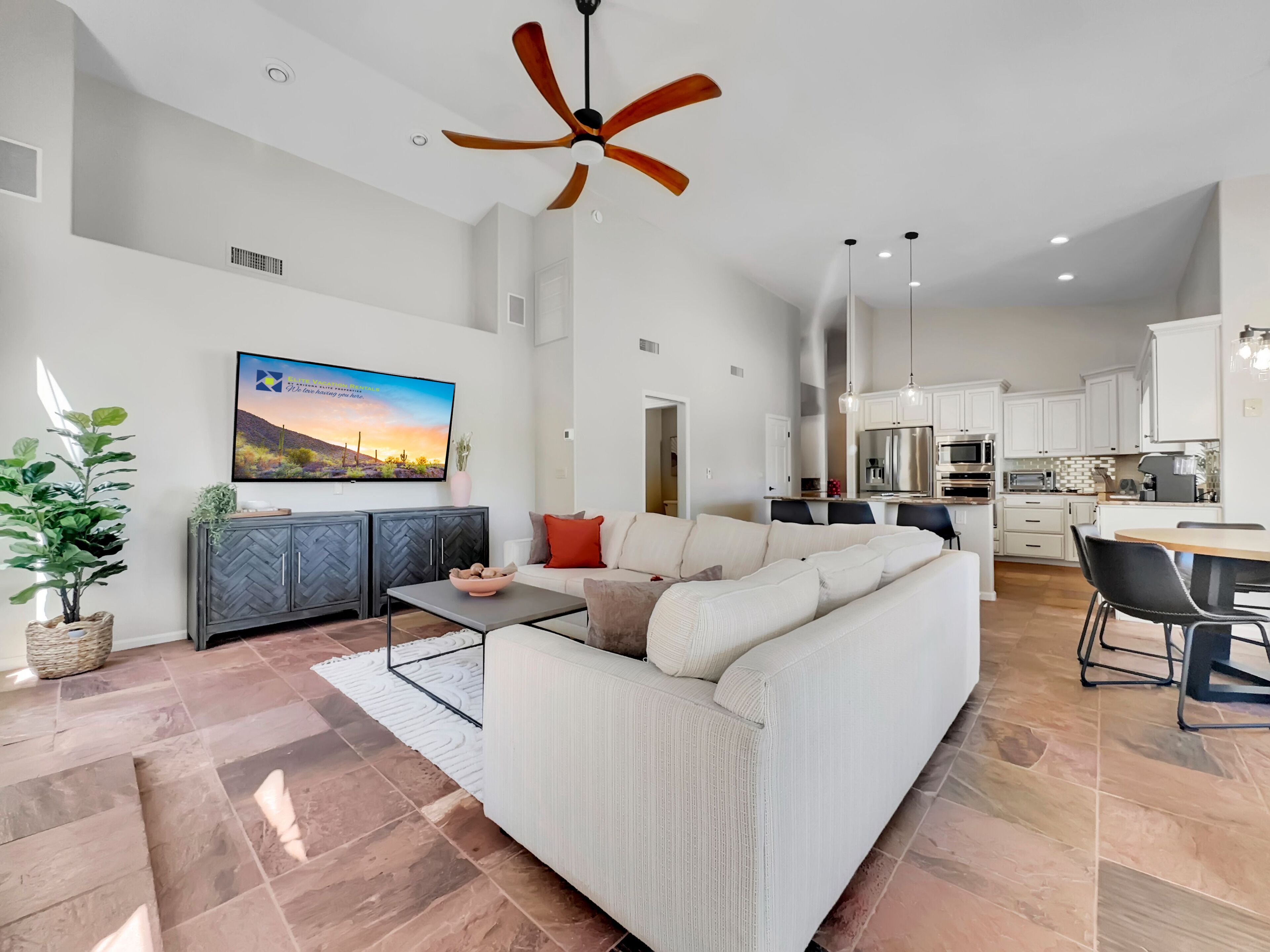 A modern living room with a white sofa, a coffee table, and a TV mounted on the wall. There is a kitchen area with white cabinets and stainless steel appliances.