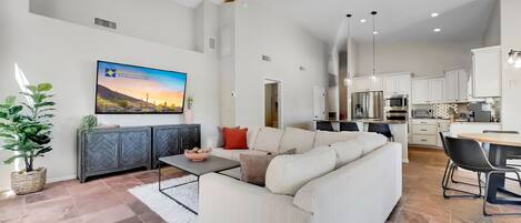 A modern living room with a white sofa, a coffee table, and a TV mounted on the wall. There is a kitchen area with white cabinets and stainless steel appliances.