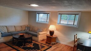 A living room with a sofa, coffee table, lamp, and a small table with a chair. There are two windows with blinds. - Private & Spacious Daylight Basement Apartment with Kitchen (Seattle)