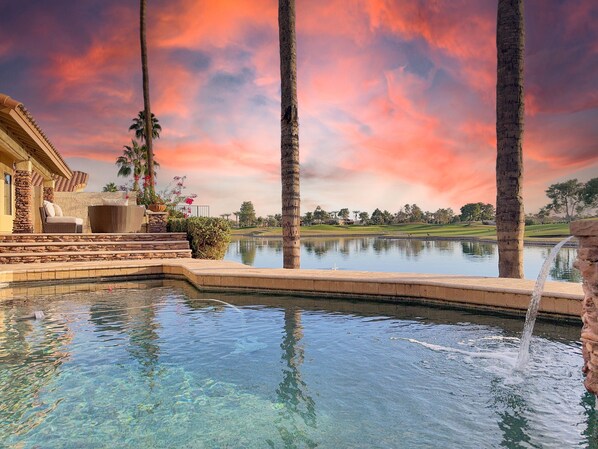 Vista despejada de una piscina con caída de agua, rodeada de palmeras y un patio de piedra con mobiliario exterior.