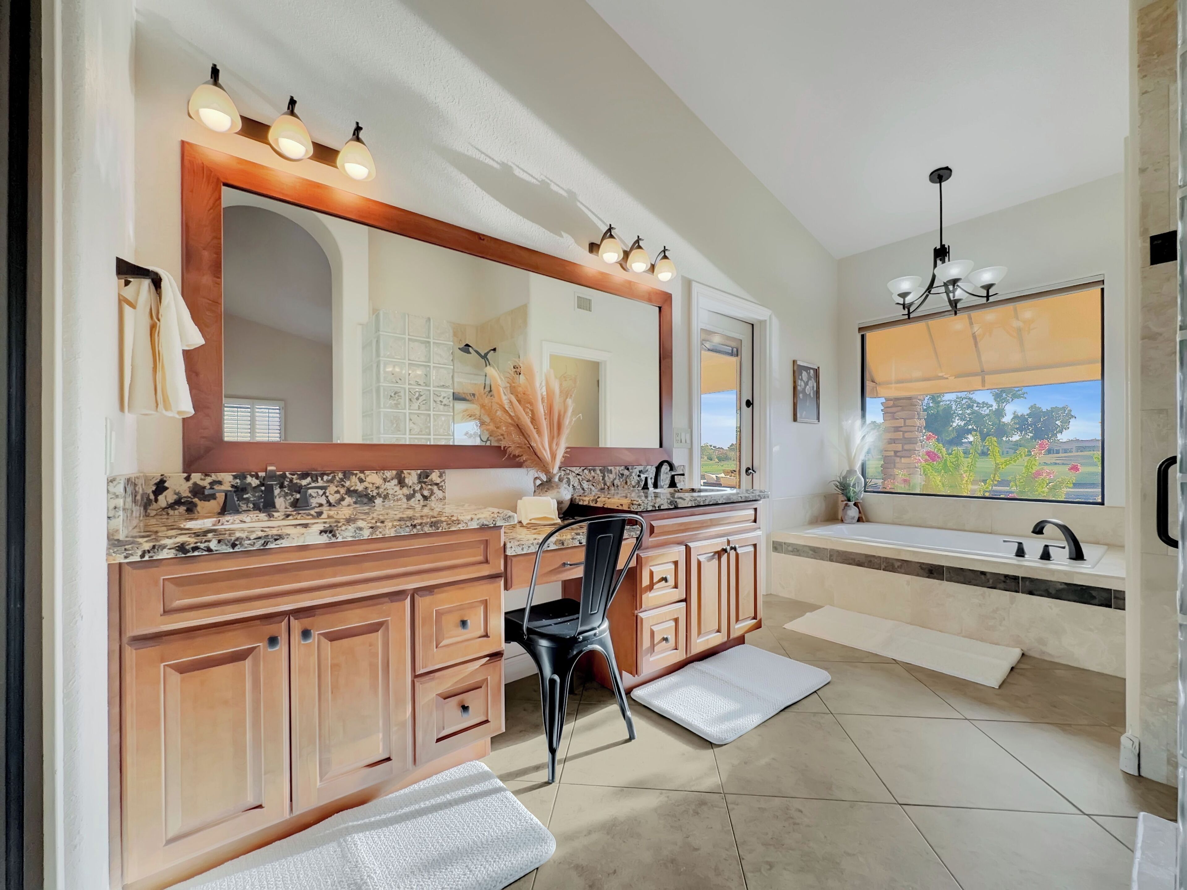 A modern bathroom with a large vanity, a mirror with lights, a sink, and a window with a view of greenery.