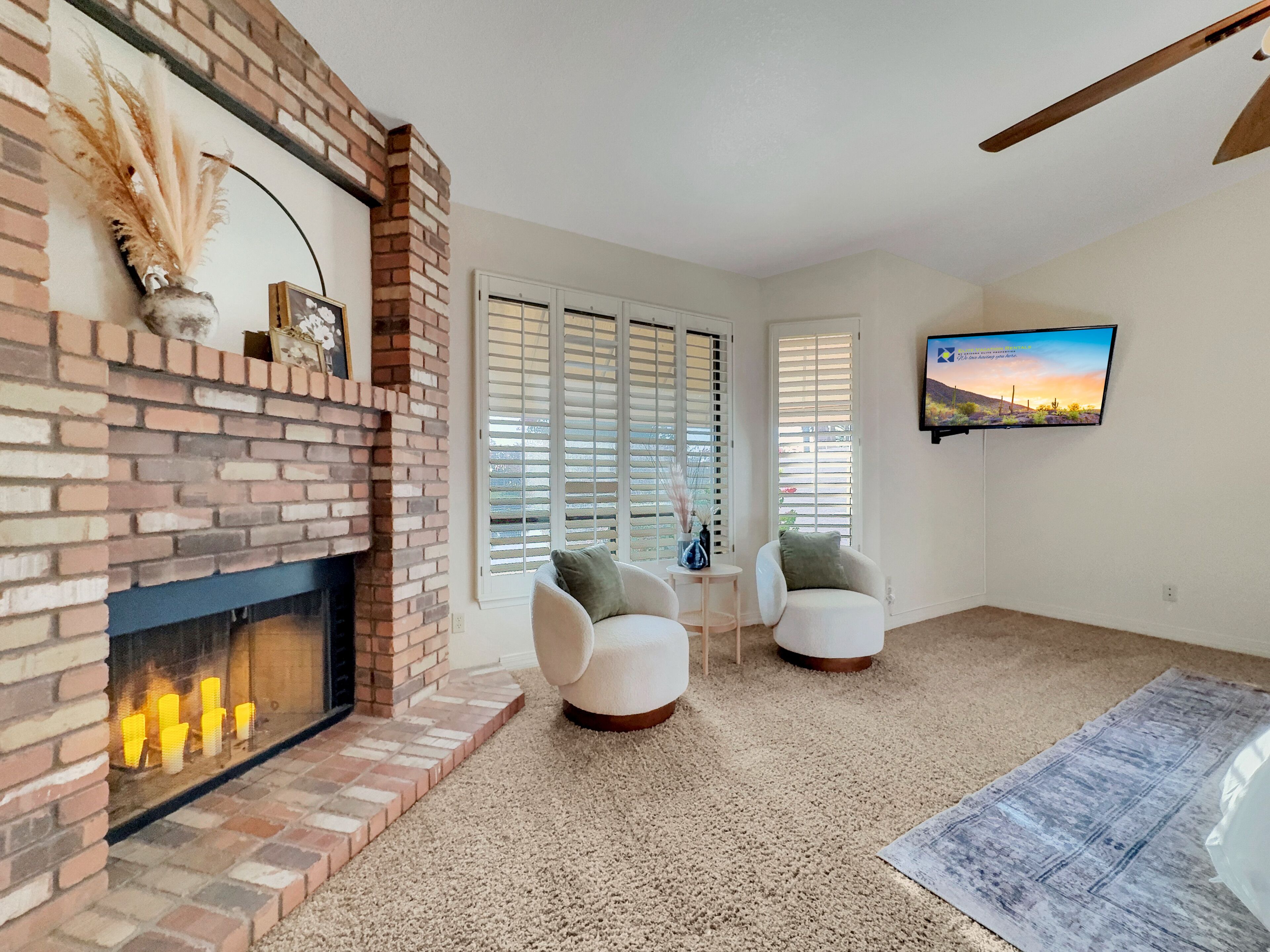 A living room with a brick fireplace, two white armchairs, a TV mounted on the wall, and a large window with blinds.
