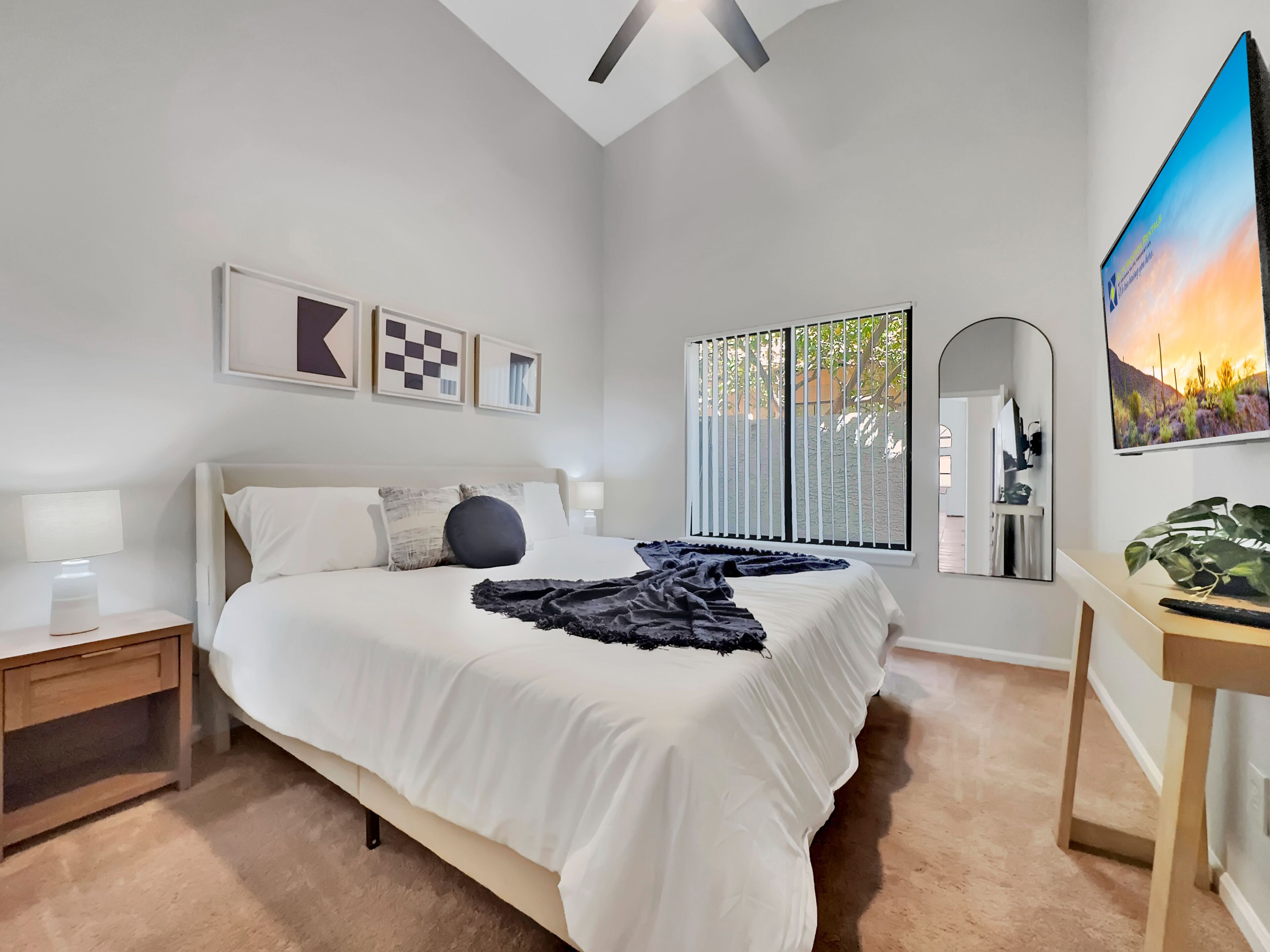 A bedroom with a bed, bedside tables, a desk, a mirror, and a window with blinds.