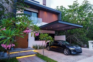 A modern house with a black car parked in front, a covered driveway, and lush greenery. - Luxury 5-Bedroom Villa in Cancun with AC, WiFi, Heated Pool and Movie Theater (Cancun)