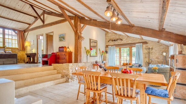 A spacious loft with a high ceiling, exposed wooden beams, a stone wall, and a dining area with wooden furniture. - La Bergerie – Charming House with Shared Pool (Le Buisson-de-Cadouin)
