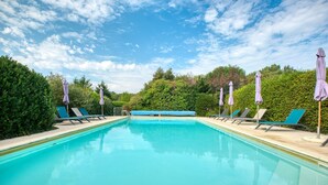 A rectangular swimming pool with blue water, surrounded by lounge chairs and umbrellas, set against a backdrop of green trees and a clear sky. - La Bergerie – Charming House with Shared Pool (Le Buisson-de-Cadouin)