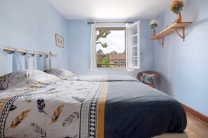 Une chambre à coucher avec un lit, une fenêtre avec des rideaux, une chaise et une étagère ornée de fleurs.
