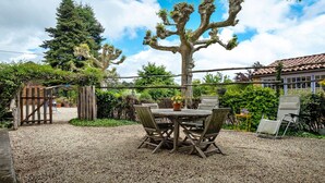 Une terrasse en gravier avec une table et des chaises en bois, entourée de verdure et d’un grand arbre.