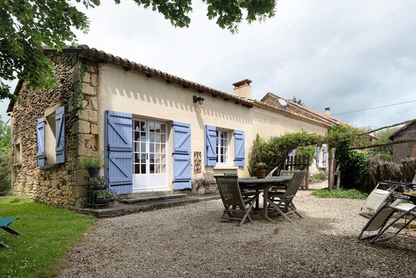 A stone building with blue shutters, a gravel driveway, and a table with chairs. - Gîte La Forge – Holiday Home with Shared Pool and Garden View in Le Buisson-de-Cadouin (Le Buisson-de-Cadouin)