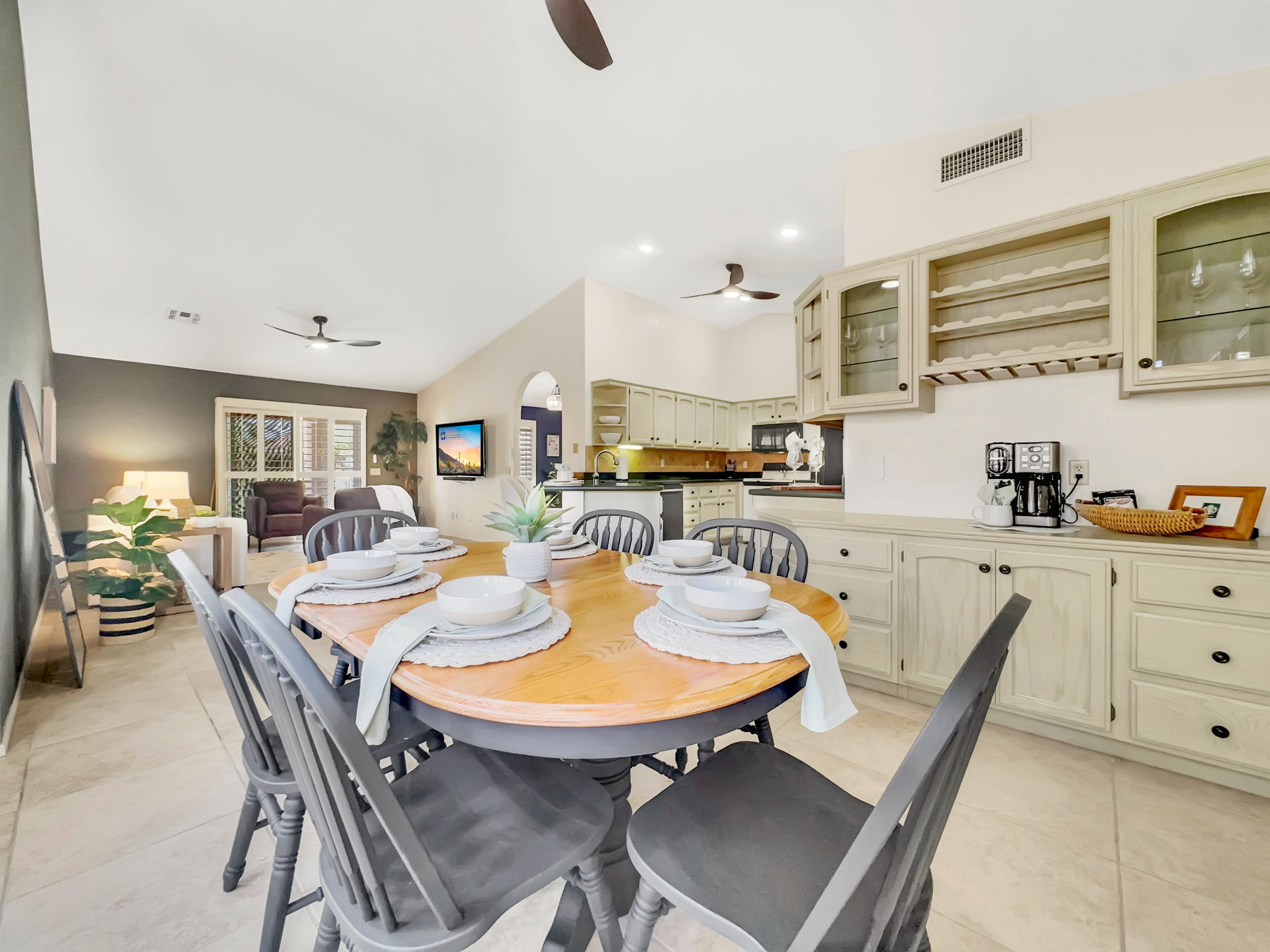 A dining area with a wooden table set for four, surrounded by chairs. There is a kitchen in the background with white cabinets and a coffee machine on the counter.