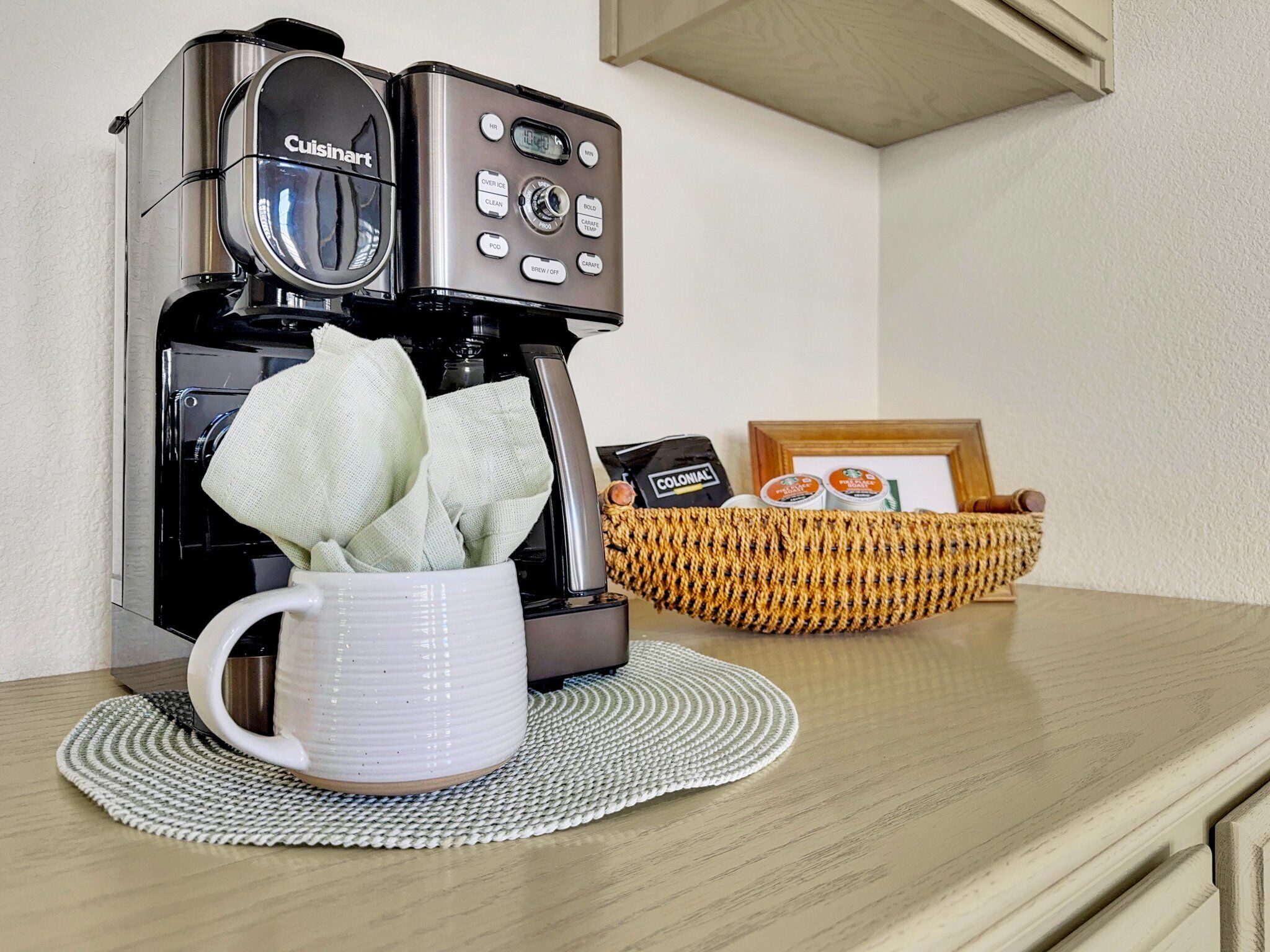 Une machine à café avec une tasse posée sur un set de table, un panier rempli d’en-cas et un tableau encadré sur une table en bois.