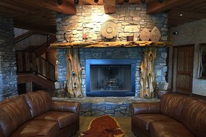 A stone fireplace with a log fire, flanked by leather sofas, and a wooden staircase in the background. - Stonewater Cove (Shell Knob)