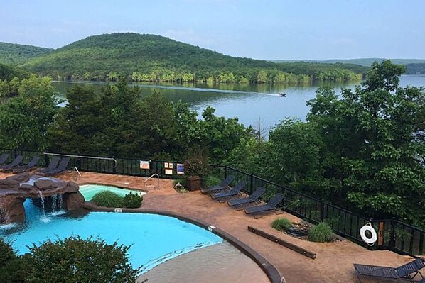 A pool area with a waterfall feature, lounge chairs, and a view of a lake surrounded by hills. - Stonewater Cove (Shell Knob)