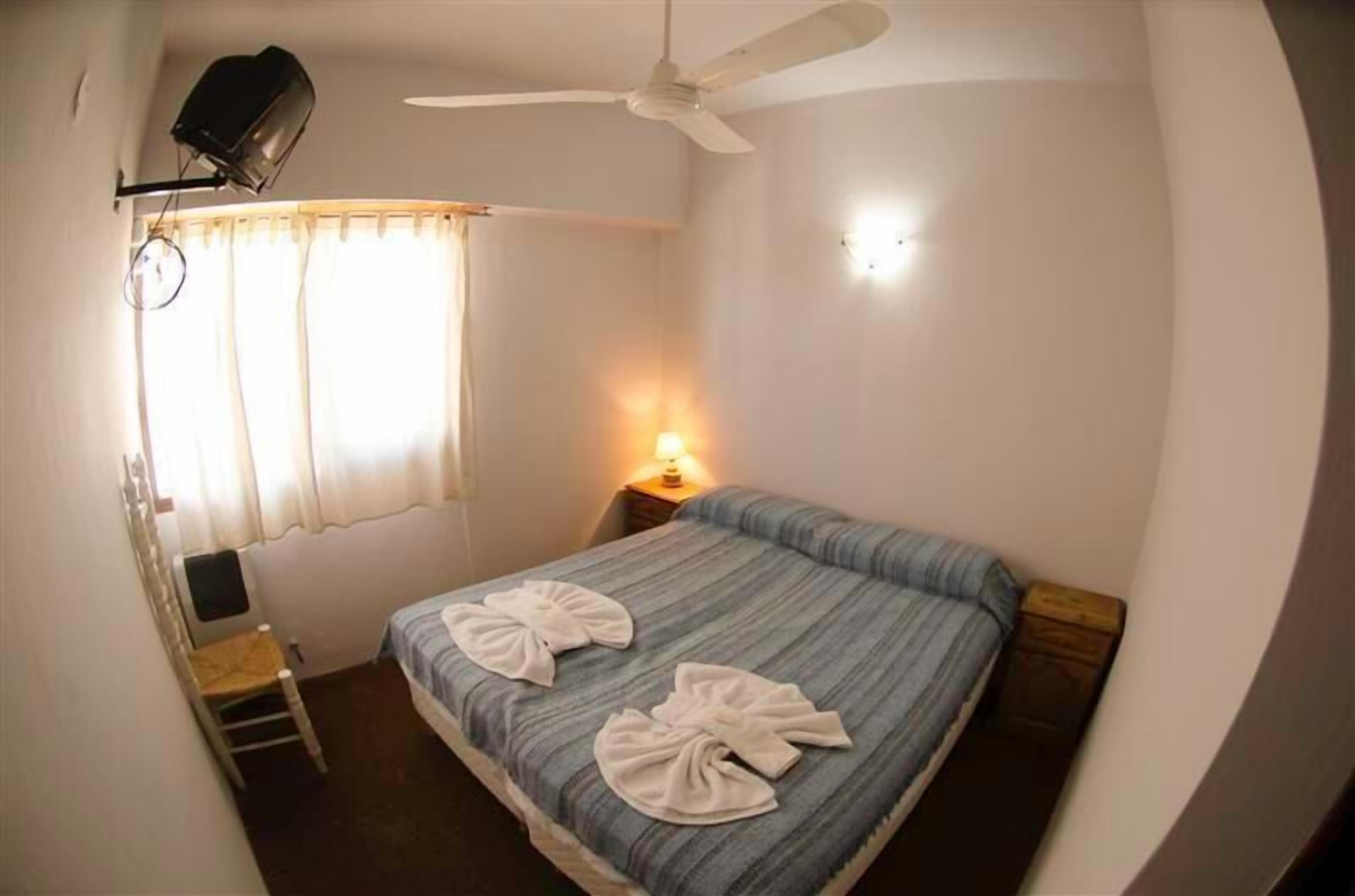 A small, single-bed room with a ceiling fan, a bedside lamp, and a window with curtains.