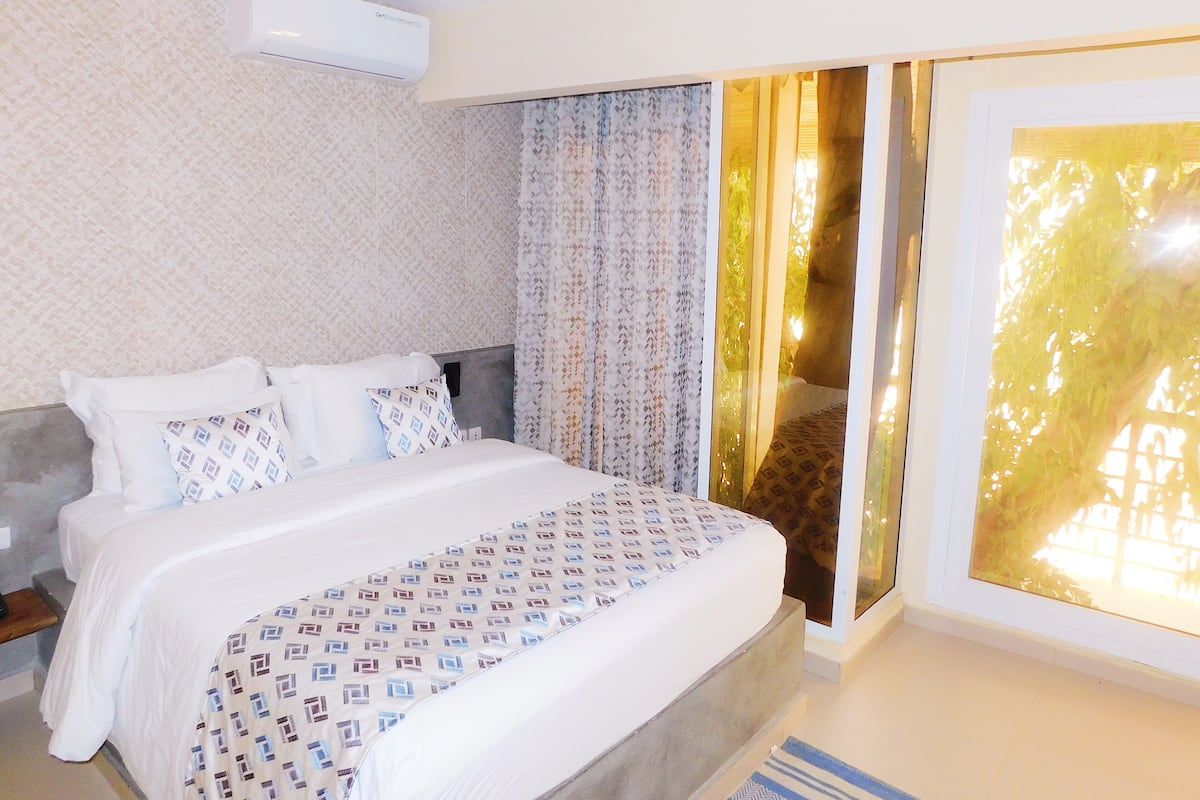 A hotel room with a bed, air conditioning unit, patterned curtains, and a view through a window.