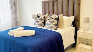 A neatly made bed with a blue bedspread and white linens, a wooden headboard, and a bedside table with a lamp.