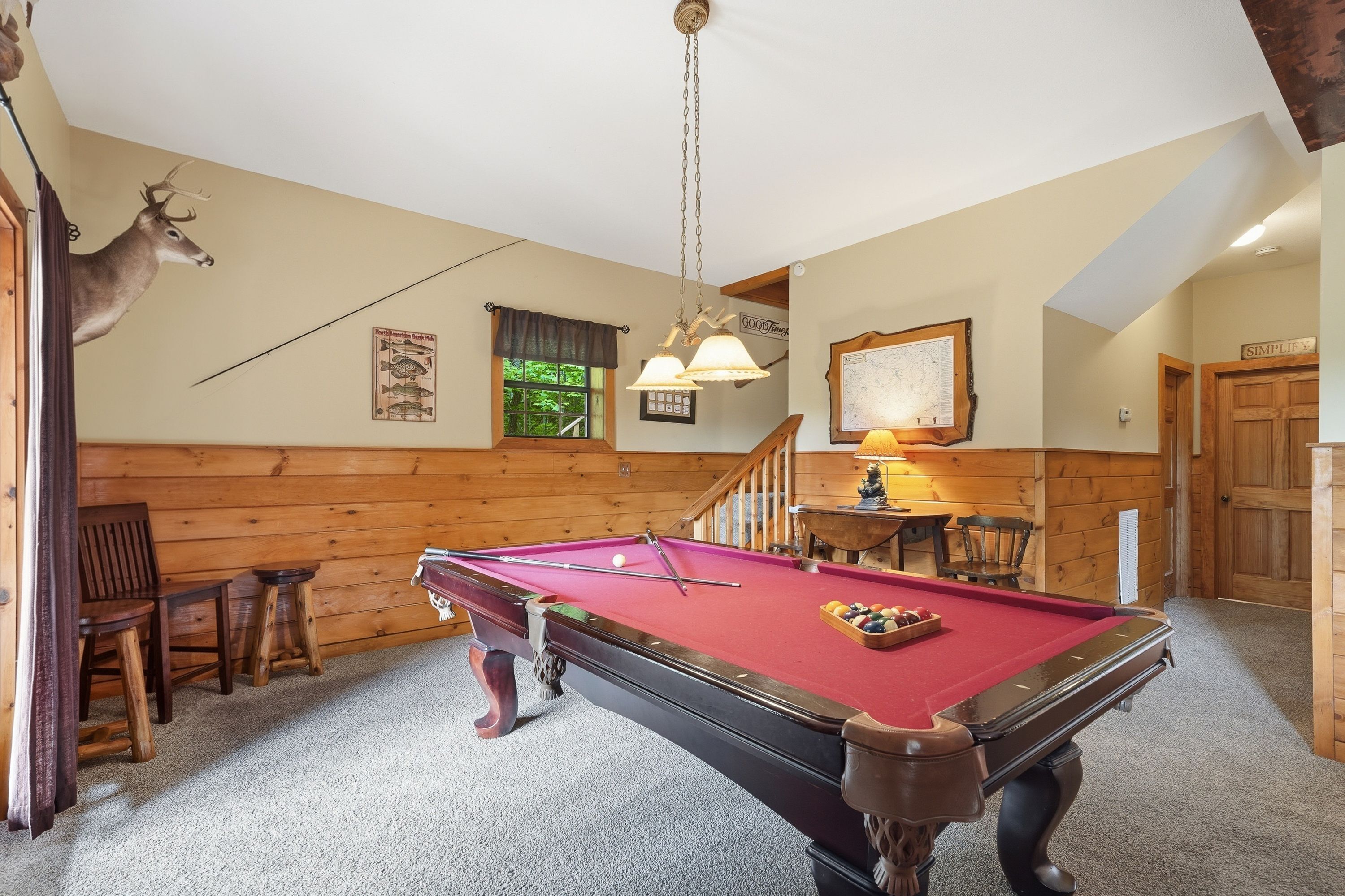 A pool table in a room with wooden walls, a mounted deer head, and a staircase.