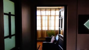 Un corridoio poco illuminato con pavimento in legno che conduce a una stanza con finestra, una sedia e un tavolino.