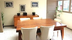 Zona pranzo con tavolo in legno e sedie imbottite bianche, quadri appesi alle pareti e una pianta in vaso.